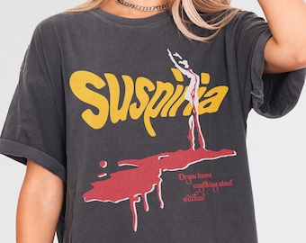 Puede incluir: Camiseta gris oscuro con la palabra "Suspiria" en amarillo, un gráfico rojo y el texto "Do you know anything about witches?" La camiseta tiene mangas enrolladas y un aspecto vintage.