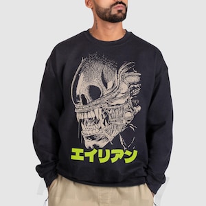 Könnte beinhalten: Schwarzes Sweatshirt mit einem detaillierten Grafikdesign eines Alien-Kreaturenkopfes in Beige und Weiß. Das japanische Wort "エイリアン" ist in Neongrün darunter gedruckt. Das Sweatshirt wird von einer Person getragen.