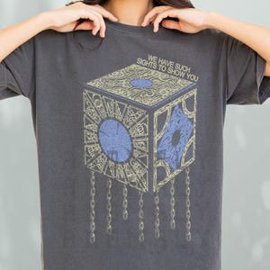 May include: Dark grey t-shirt featuring a detailed graphic of a puzzle box with blue and gold accents, suspended by chains. The text "WE HAVE SUCH SIGHTS TO SHOW YOU" is printed above the box. The shirt is a relaxed fit.