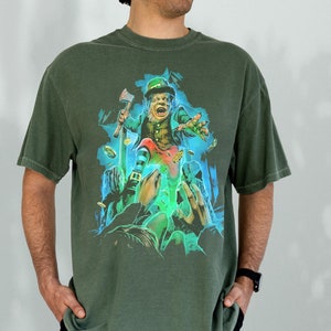 K&ouml;nnte beinhalten: Ein gr&uuml;nes T-Shirt mit einem Aufdruck eines Kobolds, der einen Topf mit Gold und eine kleine Axt h&auml;lt. Der Kobold tr&auml;gt einen gr&uuml;nen Hut und einen gr&uuml;nen Anzug. Der Aufdruck ist von einem blauen und gr&uuml;nen Schein umgeben.