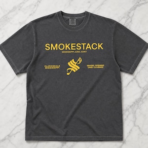 May include: Dark gray t-shirt with the text "SMOKESTACK" in yellow, above "MISSISSIPPI JUKE JOINT." Below is "CLARKSDALE MISSISSIPPI" and "GRAND OPENING (AND CLOSING)" with a yellow graphic in between. The shirt is made of a soft material.