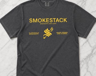 Może przedstawiać: Ciemnoszara koszulka z żółtym napisem "SMOKESTACK" nad "MISSISSIPPI JUKE JOINT". Poniżej znajdują się napisy "CLARKSDALE MISSISSIPPI" i "GRAND OPENING (AND CLOSING)" z żółtą grafiką pomiędzy nimi. Koszulka wykonana jest z miękkiego materiału.