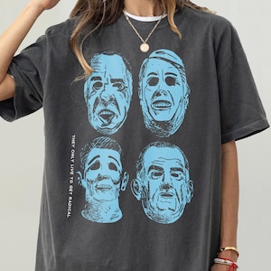May include: Dark grey t-shirt with four blue portraits. The shirt features the text "THEY ONLY LIVE TO GET RADICAL" printed vertically on the left side. The t-shirt has short sleeves and a loose fit.