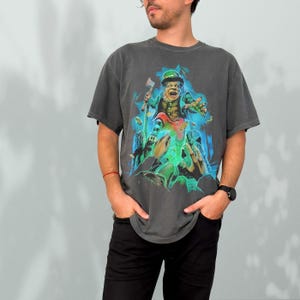 K&ouml;nnte beinhalten: Ein dunkelgraues T-Shirt mit einem grafischen Aufdruck eines gr&uuml;nen Kobolds, der eine Axt h&auml;lt und von gr&uuml;nem, leuchtendem Licht umgeben ist.
