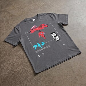 May include: Gray t-shirt featuring a graphic from the anime film Akira. The design includes a red motorcycle, a figure in a red suit, Japanese text, and a black and white illustration. The shirt is made of a soft material.