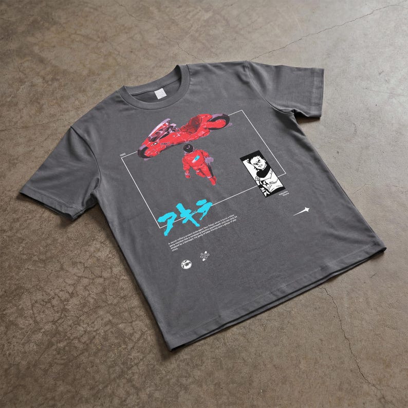 May include: Gray t-shirt featuring a graphic from the anime film Akira. The design includes a red motorcycle, a figure in a red suit, Japanese text, and a black and white illustration. The shirt is made of a soft material.