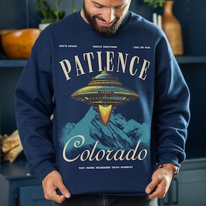 May include: Navy blue sweatshirt with a retro design featuring a UFO over a mountain range. The word "PATIENCE" is arched above the UFO, with "Colorado" below. Text includes "JOE'S DINER", "GREAT DOCTORS", "THE 39 BAR", and "WAY MORE MURDERS THAN JESSUP!"