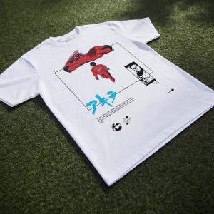 May include: White t-shirt featuring a graphic design with a red motorcycle, a figure in a red suit, and Japanese text. The design is framed by a black rectangle with additional illustrations and text. The shirt is made of a soft material.