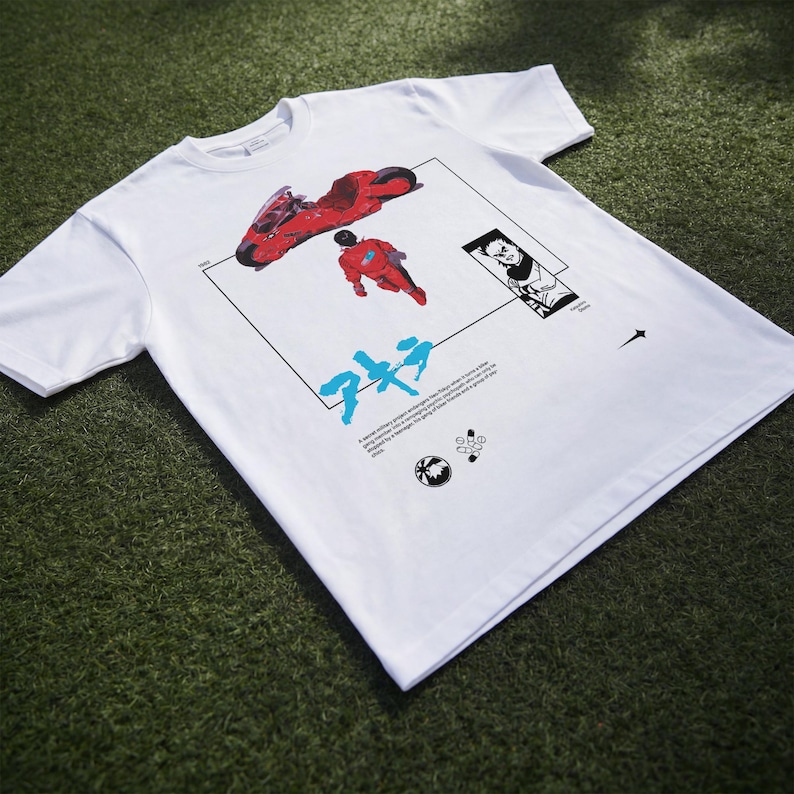 May include: White t-shirt featuring a graphic design with a red motorcycle, a figure in a red suit, and Japanese text. The design is framed by a black rectangle with additional illustrations and text. The shirt is made of a soft material.