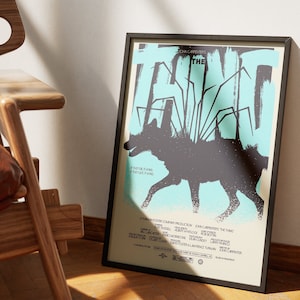 Puede incluir: Un póster enmarcado en negro con un fondo azul y blanco que presenta una silueta negra de un lobo con una criatura parecida a una araña en su espalda. El póster dice "John Carpenter's The Thing" en texto blanco.
