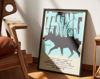 May include: A black framed poster with a blue and white background featuring a black silhouette of a wolf with a spider-like creature on its back. The poster reads "John Carpenter's The Thing" in white text.