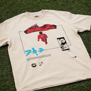 May include: Beige t-shirt featuring a graphic from the anime film Akira. The design includes a red motorcycle, a figure in a red suit, Japanese text, and a black and white illustration. The shirt also has text at the bottom.