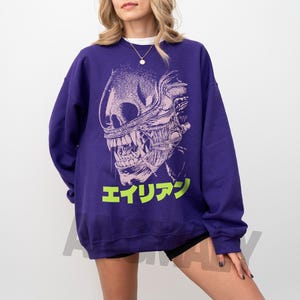 May include: A purple sweatshirt featuring a detailed, stylized illustration of an alien creature's head. The design is in white and light pink, with the Japanese word "エイリアン" in bright green at the bottom. The sweatshirt is a solid purple color.