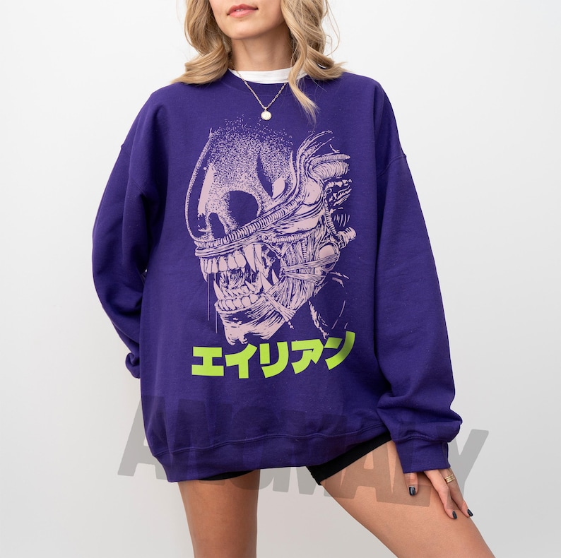 May include: A purple sweatshirt featuring a detailed, stylized illustration of an alien creature's head. The design is in white and light pink, with the Japanese word "エイリアン" in bright green at the bottom. The sweatshirt is a solid purple color.