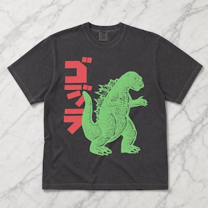 May include: A charcoal gray short-sleeve t-shirt featuring a light green graphic of a monster in a standing pose. To the left of the monster, there is red Japanese text arranged vertically. The shirt has a crew neckline and a standard fit.
