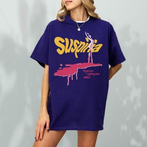 May include: A navy blue t-shirt with the word "Suspiria" in yellow, and a pink graphic of a figure with the text "Do you know anything about witches?" The shirt has short sleeves and a crew neck.