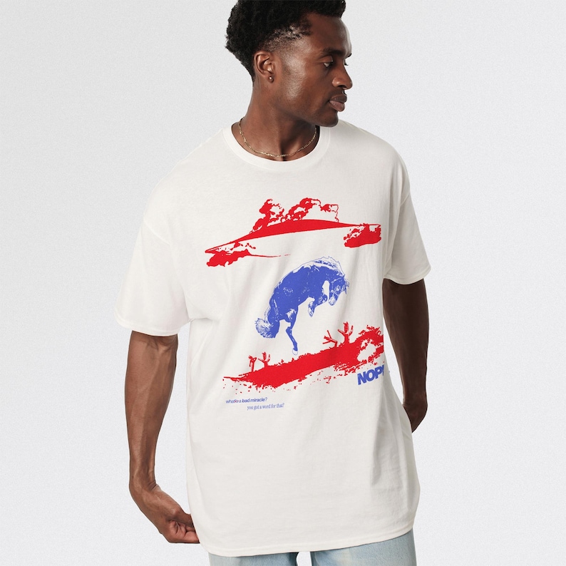 May include: White t-shirt with a graphic design featuring a blue bucking bronco and red abstract shapes. The word "NOP" is printed in blue, and smaller text is visible below the design.