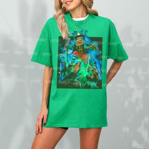 K&ouml;nnte beinhalten: Gr&uuml;nes T-Shirt mit einem Aufdruck eines Kobolds, der einen Topf mit Gold und einen Kn&uuml;ppel h&auml;lt. Der Kobold ist von anderen Figuren umgeben.