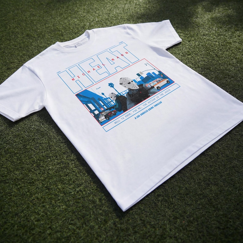 May include: White t-shirt featuring a graphic from the movie "Heat". The design includes the word "HEAT" in blue and red, with the name "MICHAEL MANN" below. The graphic depicts a scene with cars and figures, with the text "WELL YA KNOW, FOR ME, THE ACTION IS THE JUICE. A LOS ANGELES CRIME THRILLER."