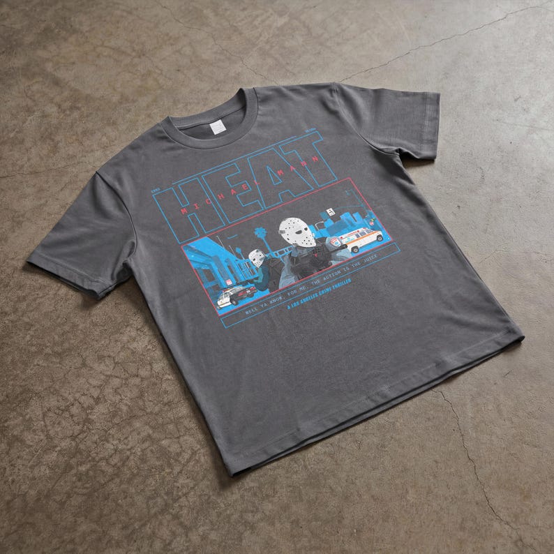 May include: Dark gray t-shirt featuring a graphic design with the word "HEAT" in blue and a scene with figures wearing hockey masks. The graphic also includes the text "MICHAEL MANN" and other text.