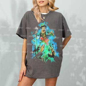 K&ouml;nnte beinhalten: Ein dunkelgraues T-Shirt mit einem grafischen Aufdruck eines Kobolds, der einen Topf mit Gold und eine Axt h&auml;lt. Der Kobold tr&auml;gt einen gr&uuml;nen Hut und eine gr&uuml;ne Weste. Die Grafik ist von gr&uuml;nen und blauen Wirbeln umgeben.