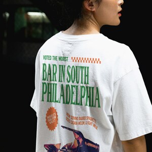 Puede incluir: Camiseta blanca con un estampado gr&aacute;fico verde y naranja que dice "Voted the worst Bar in South Philadelphia". El gr&aacute;fico tambi&eacute;n incluye un dibujo de una botella rota y el texto "Novelty Egg for Sale", "Now serving bagged spaghetti, used casaba melon & fight milk", y "Proud distributor of Frank's Fluids Paddy's Irish Pub".