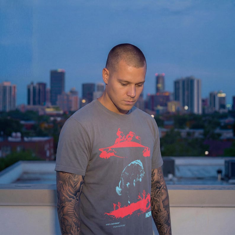 May include: A gray t-shirt with a red and blue graphic design. The design appears to be an abstract image. The person wearing the shirt is standing on a rooftop with a city skyline in the background.