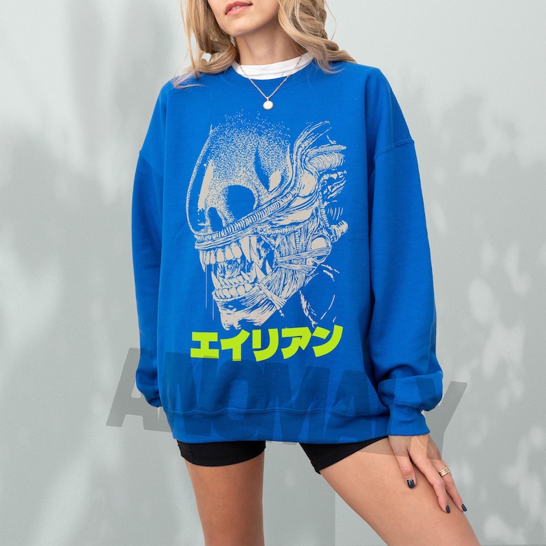 May include: Royal blue sweatshirt featuring a detailed, off-white graphic of an alien creature's head. The design includes sharp teeth and intricate skeletal details. Below the graphic is Japanese text in neon yellow.