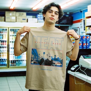 May include: Tan t-shirt featuring the word "HEAT" in blue and red, with "MICHAEL MANN" below. The graphic depicts a scene with figures wearing masks, cars, and an ambulance. Text at the bottom reads "WELL TO FEEL FOR ME, THE ACTION IS THE JUICE".