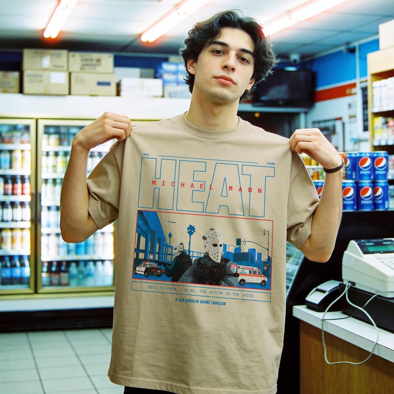 May include: Tan t-shirt featuring the word "HEAT" in blue and red, with "MICHAEL MANN" below. The graphic depicts a scene with figures wearing masks, cars, and an ambulance. Text at the bottom reads "WELL TO FEEL FOR ME, THE ACTION IS THE JUICE".