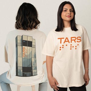 May include: Off-white t-shirts with the word "TARS" in orange, along with braille dots. One shirt features a building graphic on the back. The shirts are short-sleeved and have a relaxed fit.