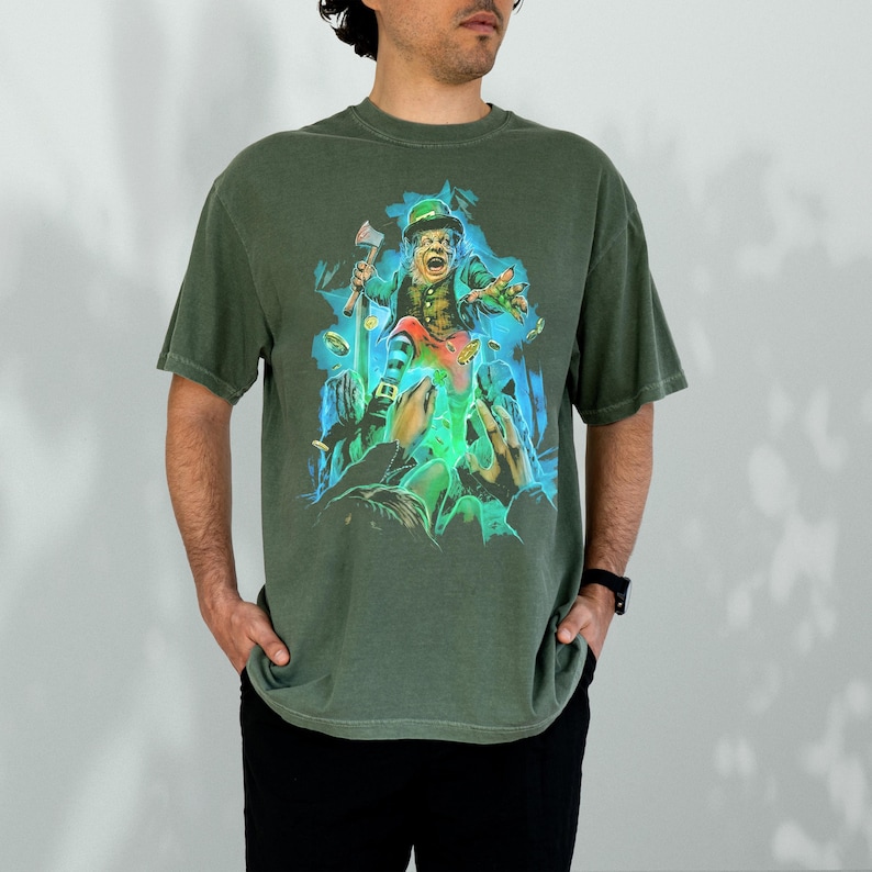 K&ouml;nnte beinhalten: Ein gr&uuml;nes T-Shirt mit einem Aufdruck eines Kobolds, der einen Topf mit Gold und eine kleine Axt h&auml;lt. Der Kobold tr&auml;gt einen gr&uuml;nen Hut und einen gr&uuml;nen Anzug. Der Aufdruck ist von einem blauen und gr&uuml;nen Schein umgeben.