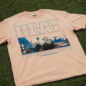 May include: A light peach-colored t-shirt featuring a graphic from the movie "Heat." The design includes the word "HEAT" in blue block letters, the name "MICHAEL MANN," and a scene with masked figures and vehicles. The text "WELL YA KNOW, FOR ME, THE ACTION IS THE JUICE" is also included.