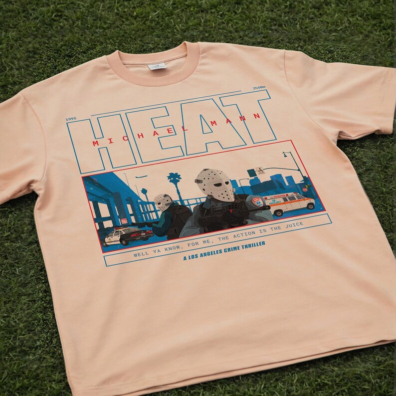 May include: A light peach-colored t-shirt featuring a graphic from the movie "Heat." The design includes the word "HEAT" in blue block letters, the name "MICHAEL MANN," and a scene with masked figures and vehicles. The text "WELL YA KNOW, FOR ME, THE ACTION IS THE JUICE" is also included.