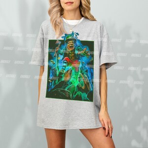 K&ouml;nnte beinhalten: Ein graues T-Shirt mit einem grafischen Aufdruck eines gr&uuml;nen Kobolds, der einen goldenen Topf mit M&uuml;nzen h&auml;lt. Der Kobold tr&auml;gt einen gr&uuml;nen Hut und einen gr&uuml;nen Anzug. Der Hintergrund ist dunkelgr&uuml;n und blau.