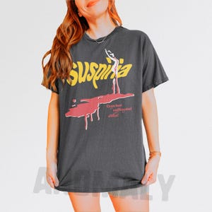 May include: Dark gray t-shirt featuring the word "Suspiria" in yellow, with a red graphic of a dancer and the text "Do you know anything about witches?" A vintage-style tee.