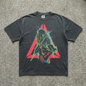 Puede incluir: Camiseta gris oscuro con un gráfico colorido de un personaje Predator dentro de un triángulo rojo. El diseño incluye rayos y un Predator sosteniendo un arma. La camiseta es de manga corta y tiene cuello redondo.