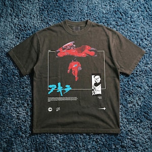 May include: Dark gray t-shirt featuring a red motorcycle and a figure in a red suit, with Japanese text and illustrations. The shirt has a vintage look and feel, with a crew neck and short sleeves.
