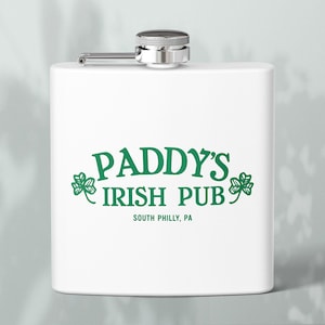 Könnte beinhalten: Weißer Flachmann mit silbernem Verschluss und Scharnier. Auf der Vorderseite steht in grüner Schrift "PADDY'S IRISH PUB" mit Kleeblatt-Akzenten und dem Standort "SOUTH PHILLY, PA".