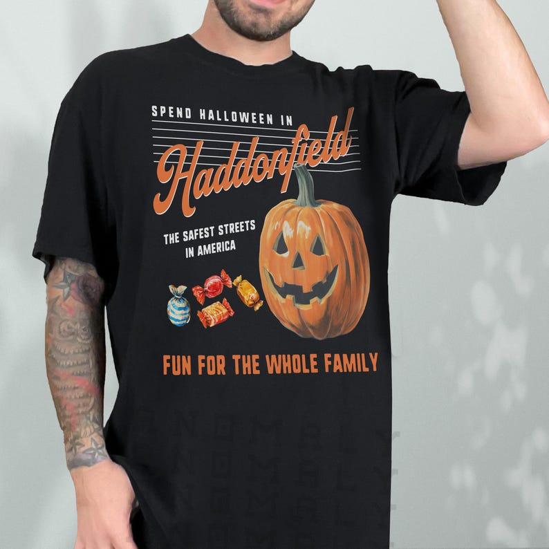May include: Black t-shirt with a Halloween-themed graphic. The design features a carved orange pumpkin, candy, and text that reads "Spend Halloween in Haddonfield, The Safest Streets in America, Fun for the Whole Family."