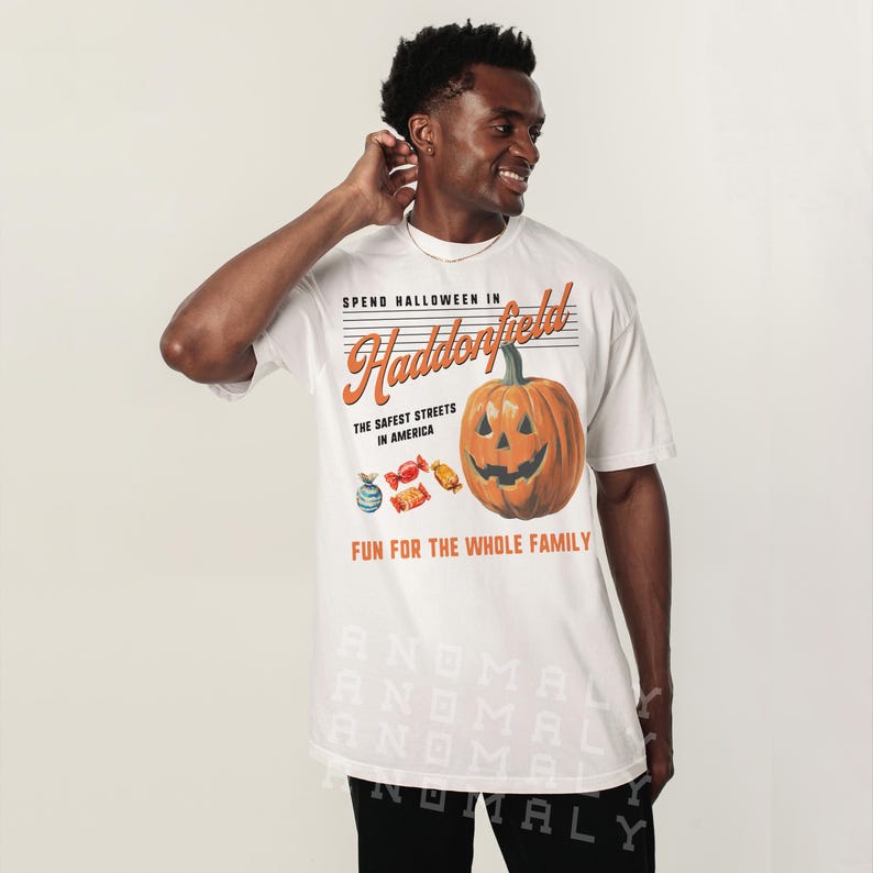 May include: White t-shirt with a Halloween-themed graphic. The design features a smiling jack-o'-lantern, candy, and the text "SPEND HALLOWEEN IN Haddonfield, The Safest Streets in America, FUN FOR THE WHOLE FAMILY."