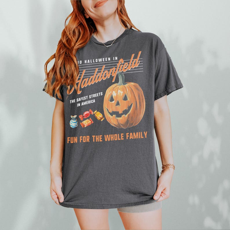 May include: Dark gray t-shirt with a Halloween-themed graphic. The design features a smiling jack-o'-lantern, candy, and text that reads "10 Halloween in Haddonfield, The Safest Streets in America, Fun for the Whole Family."