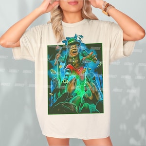 K&ouml;nnte beinhalten: Ein wei&szlig;es T-Shirt mit einem grafischen Aufdruck eines gr&uuml;nen Kobolds, der einen Topf mit Gold h&auml;lt. Der Kobold tr&auml;gt einen gr&uuml;nen Hut und einen gr&uuml;nen Anzug. Der Hintergrund ist ein dunkler gr&uuml;ner Wald.