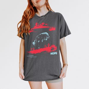 May include: Dark grey t-shirt with a red and blue graphic design. The design includes a stylized animal figure and the word "NOPE" in blue. The shirt also has the text "what's a bad miracle? you gotta want the thing!"