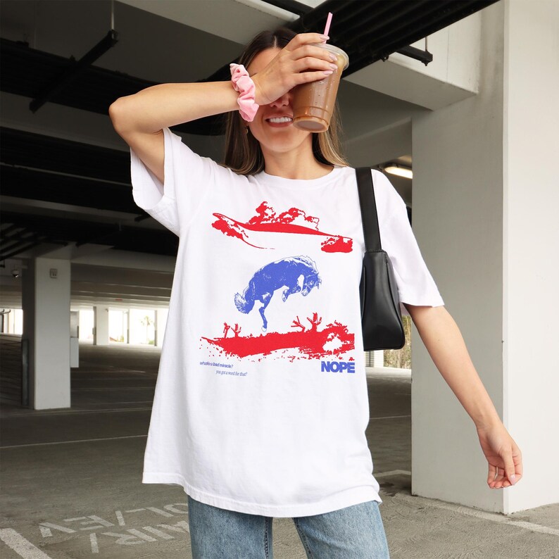 May include: White t-shirt with a graphic design featuring red and blue abstract shapes and the word "NOPE" in blue. The shirt has a crew neck and short sleeves. The design includes text at the bottom. The person is holding a drink.