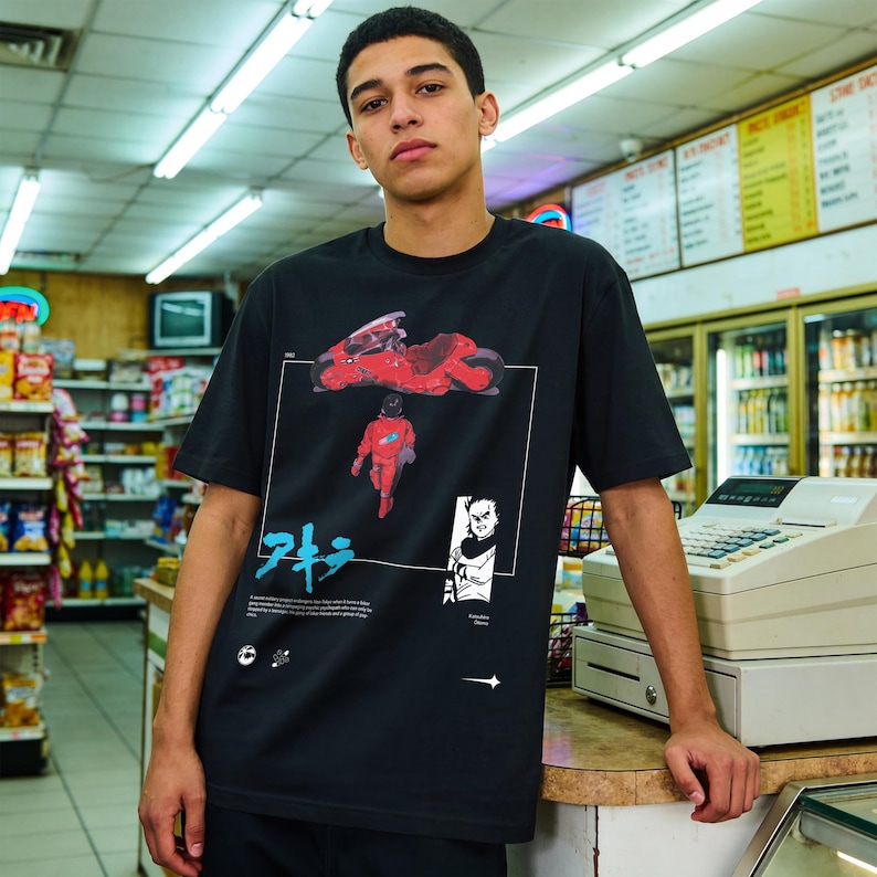May include: Black t-shirt featuring a red motorcycle and a figure in a red suit, with Japanese text and other graphics. The design is framed by a white border. The shirt is worn by a person in a convenience store.