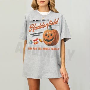 May include: A gray t-shirt with a Halloween-themed graphic. The design features a jack-o'-lantern, candy, and the text "SPEND HALLOWEEN IN Haddonfield, THE SAFEST STREETS IN AMERICA, FUN FOR THE WHOLE FAMILY."