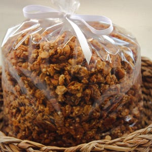 May include: A cellophane bag of granola, tied with a white ribbon, rests in a woven basket. The granola is a mix of golden brown and tan pieces. The basket is made of woven, light brown material.
