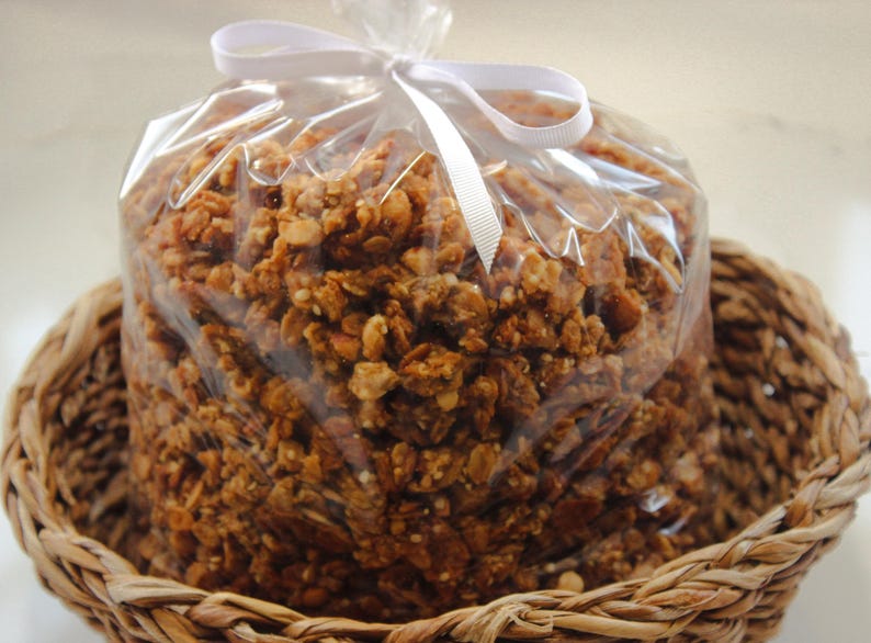May include: A cellophane bag of granola, tied with a white ribbon, rests in a woven basket. The granola is a mix of golden brown and tan pieces. The basket is made of woven, light brown material.