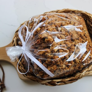 May include: A clear plastic bag of granola, tied with a white ribbon, is presented in a woven basket. The basket sits on a wooden chopping board with a handle. The granola is a mix of golden brown and tan ingredients.